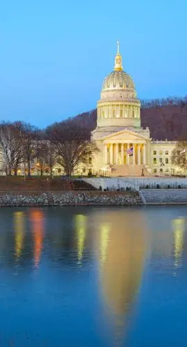 A grand, illuminated capitol building at twilight reflects elegantly on the calm waters of a nearby river.