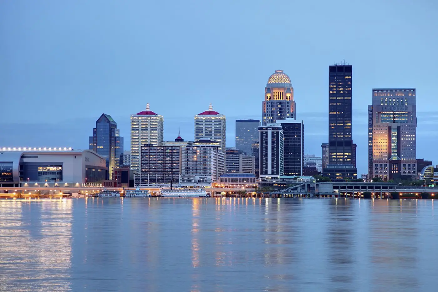 A skyline of a modern city with illuminated buildings reflected in a large body of water during twilight.