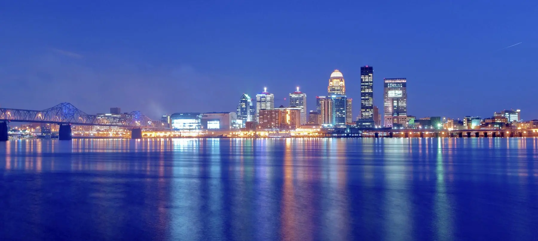 A panoramic night view of a city skyline with illuminated buildings reflected on a large body of water.