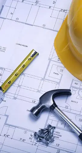 A yellow construction helmet, a tape measure, a hammer, and screws are placed on top of architectural blueprints.