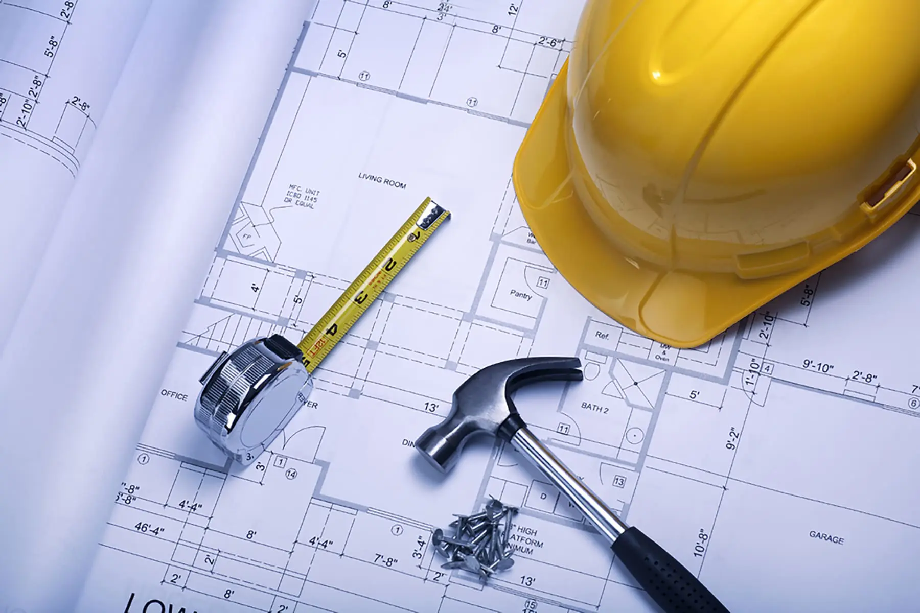 A yellow construction helmet, a tape measure, a hammer, and screws are placed on top of architectural blueprints.