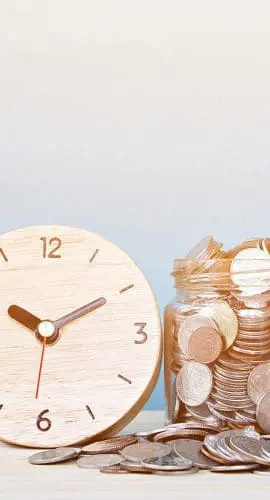 A wooden clock is placed next to a jar filled with coins, with more coins scattered around the jar.