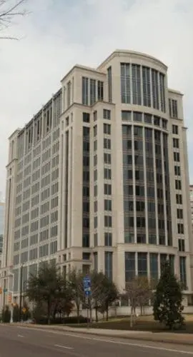 A wide street is lined with tall, modern buildings and trees, leading to a large corner building with a rounded design element on top.