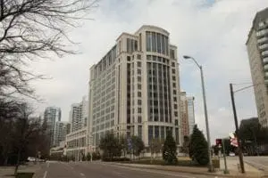 A wide street is lined with tall, modern buildings and trees, leading to a large corner building with a rounded design element on top.