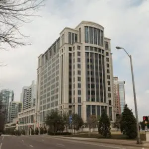 A wide street is lined with tall, modern buildings and trees, leading to a large corner building with a rounded design element on top.