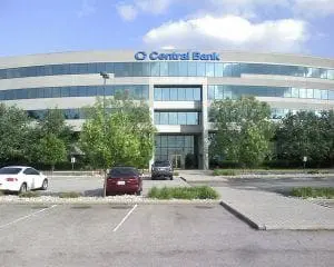 A modern multi-story building with a "Central Bank" sign, surrounded by a parking lot with several parked cars and trees.