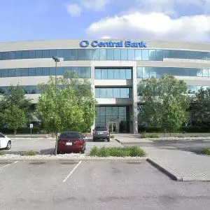 A modern multi-story building with a "Central Bank" sign, surrounded by a parking lot with several parked cars and trees.