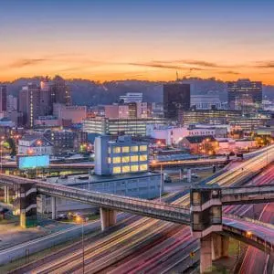 A vibrant cityscape is captured at dusk, featuring illuminated buildings and intersecting highways against a backdrop of rolling hills and a colorful sky.