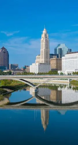 A city skyline with modern buildings is reflected in a calm river under a blue sky.