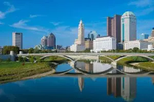 A city skyline with modern buildings is reflected in a calm river under a blue sky.