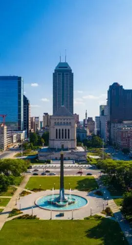 Aerial view of a city skyline with a park and a prominent fountain in the foreground under a clear blue sky.