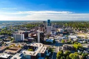 An aerial view of a downtown district showcases a mix of high-rise buildings, urban streets, and scattered greenery under a blue sky with light cloud cover.