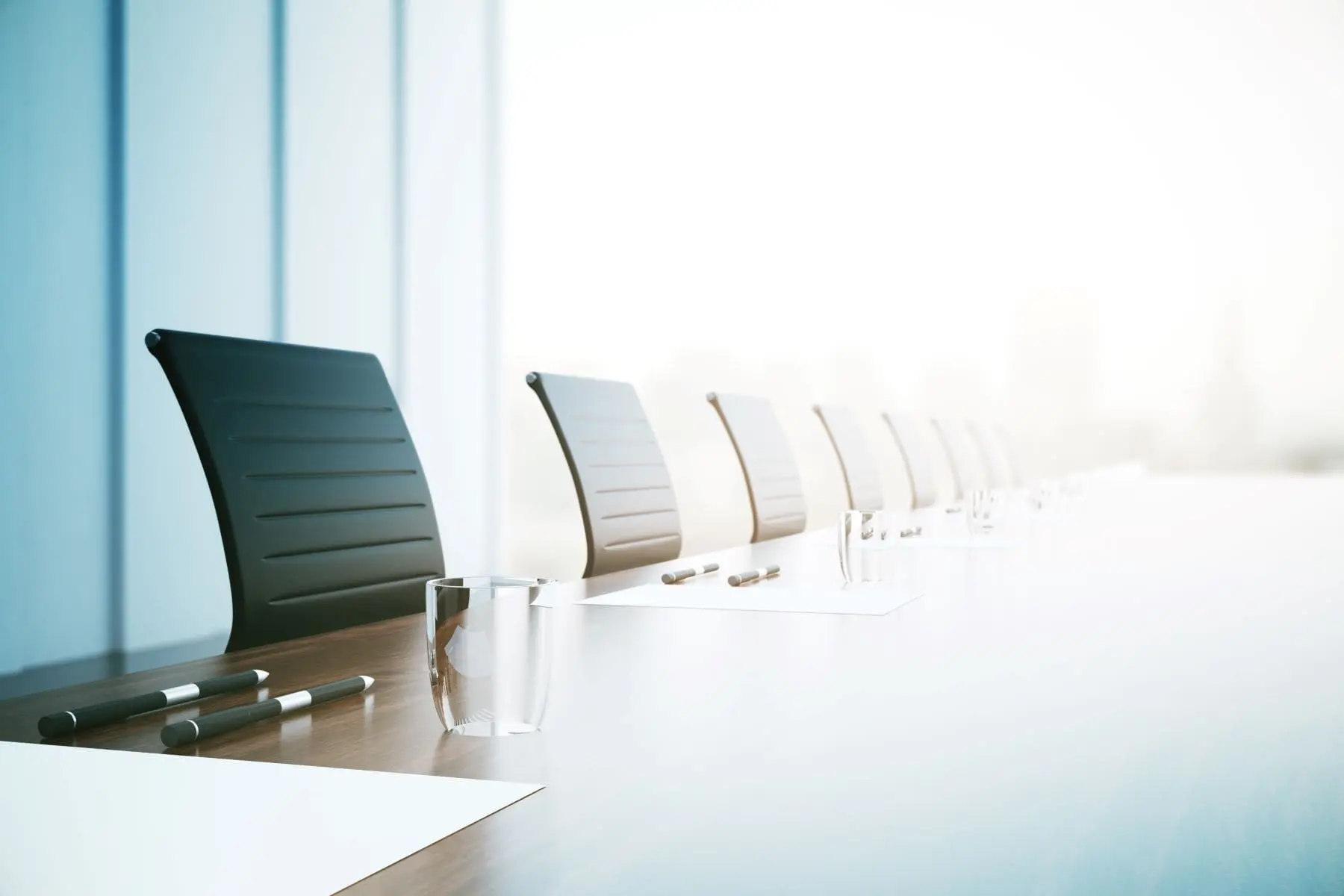 A row of black chairs alongside a modern conference table with a glass of water, indicating a professional or business setting, with bright natural light in the background.