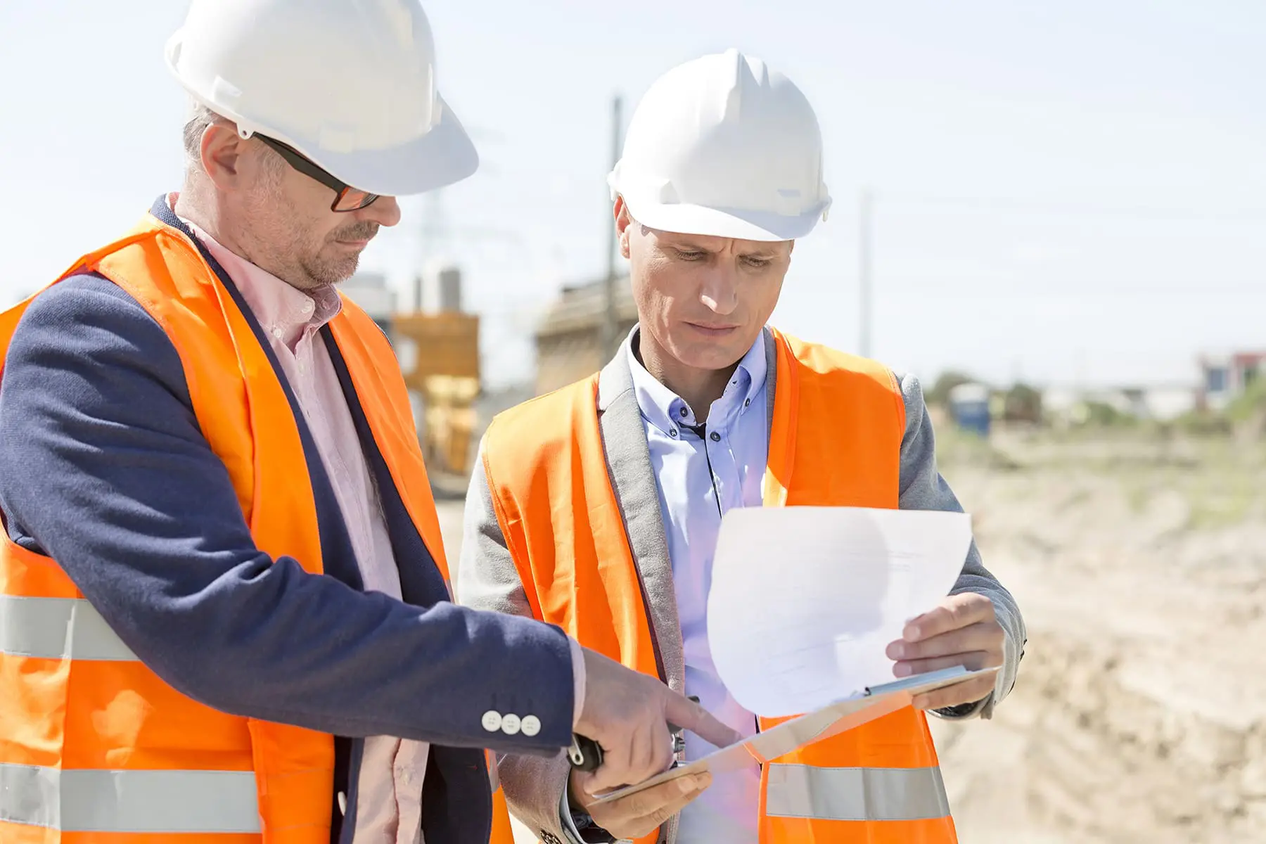 Two individuals wearing hard hats and high-visibility vests are examining a document at a construction site.