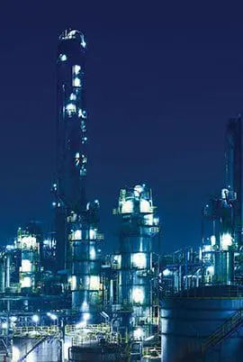 A nighttime scene of an illuminated industrial complex with multiple structures and bright lights set against a dark blue sky.