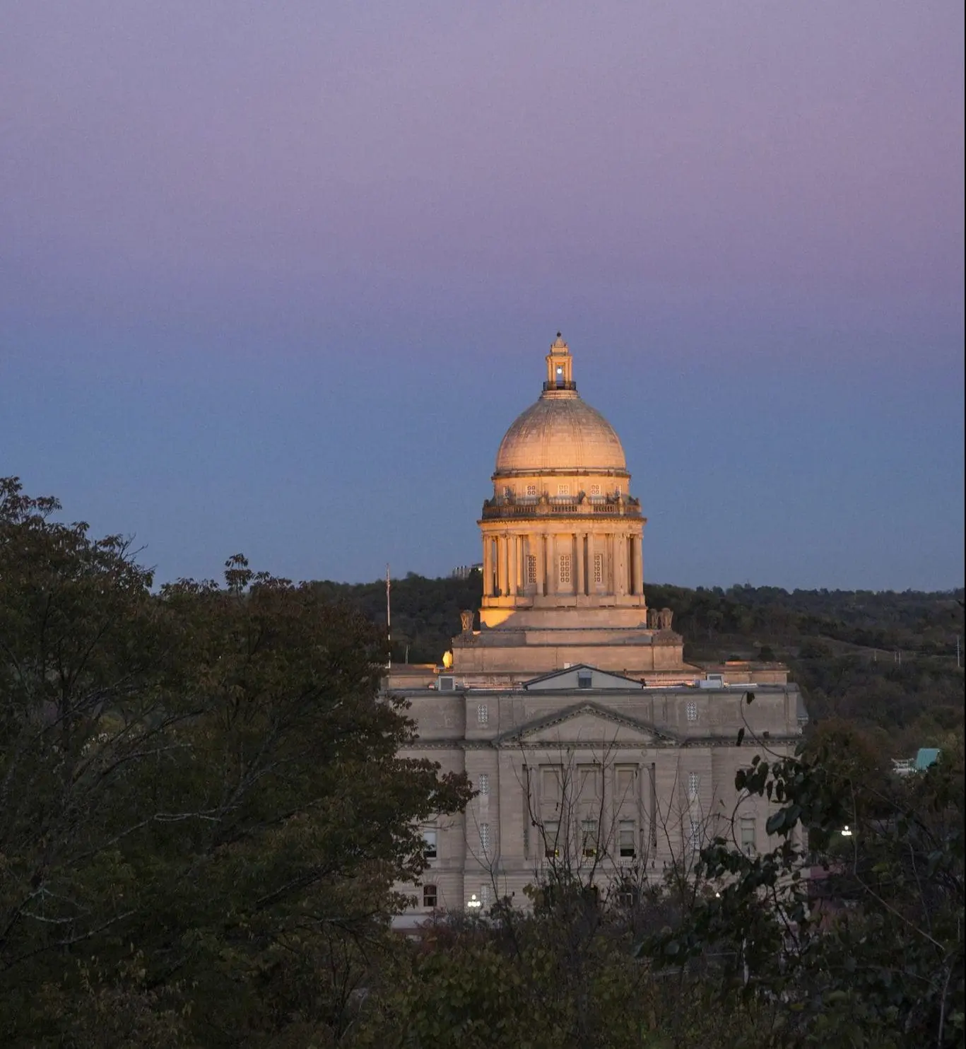 A grand building with a prominent dome is illuminated against a twilight sky, surrounded by trees.