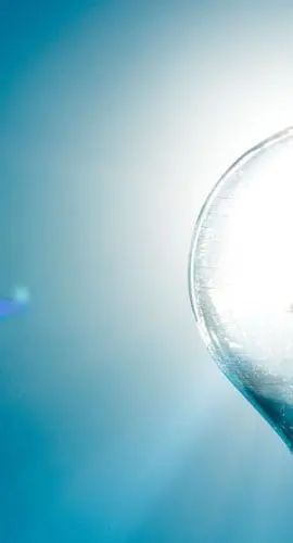 A clear light bulb is illuminated against a bright blue background with light flares.