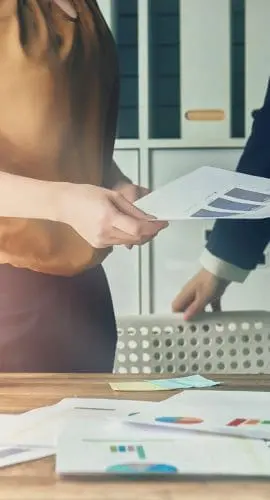 Two professionals are exchanging documents over a desk strewn with charts and a laptop, illuminated by warm sunlight.