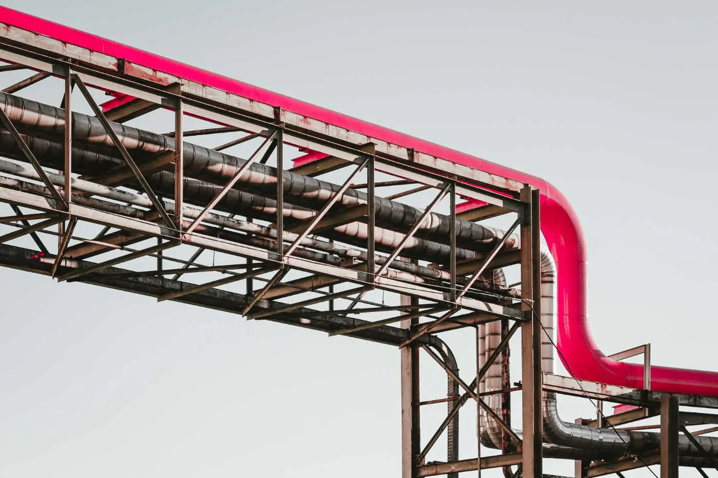 A red framework encloses a series of industrial pipes against a pale sky.