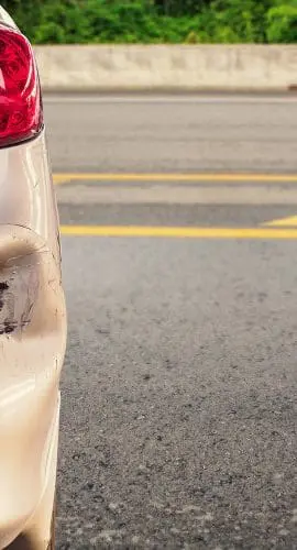 A silver car with a dented bumper is parked beside a road with a yellow dividing line.