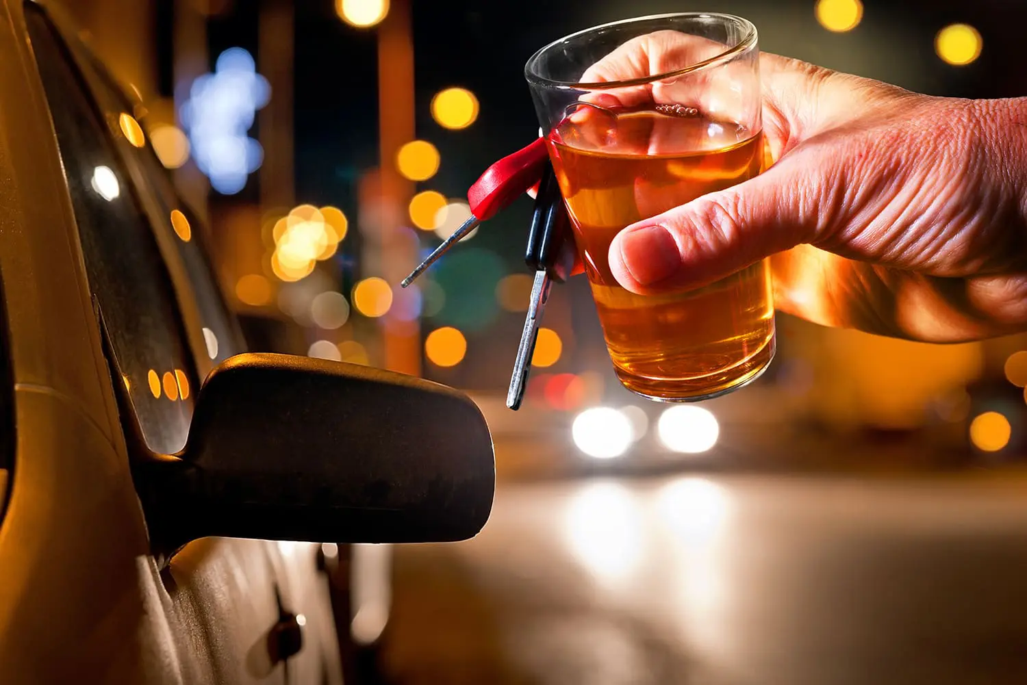 A hand is holding a glass of alcohol and car keys against a backdrop of blurred city lights at night.