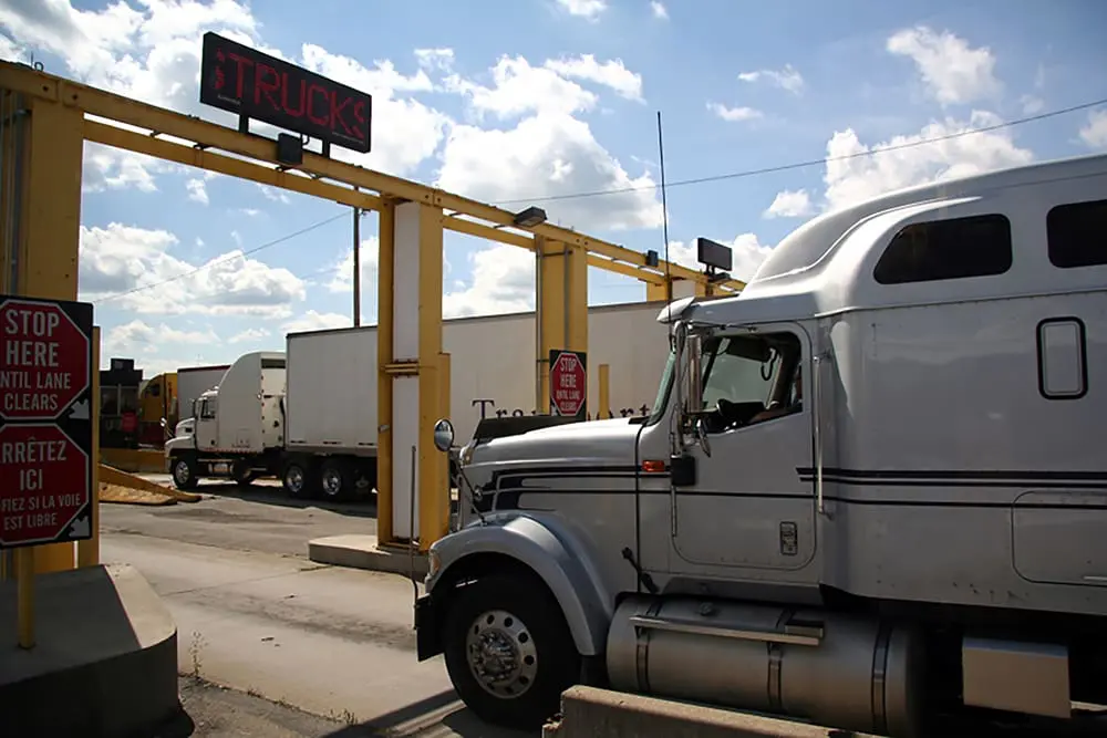 A semi-truck is passing through a commercial vehicle checkpoint with various traffic signs and another truck visible in the background.