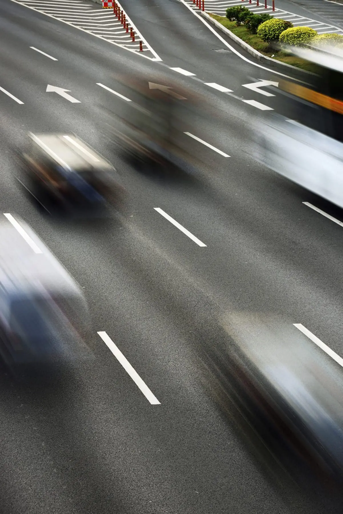 Vehicles captured in motion blur travel along a multi-lane road with marked lanes and a central barrier with shrubbery.