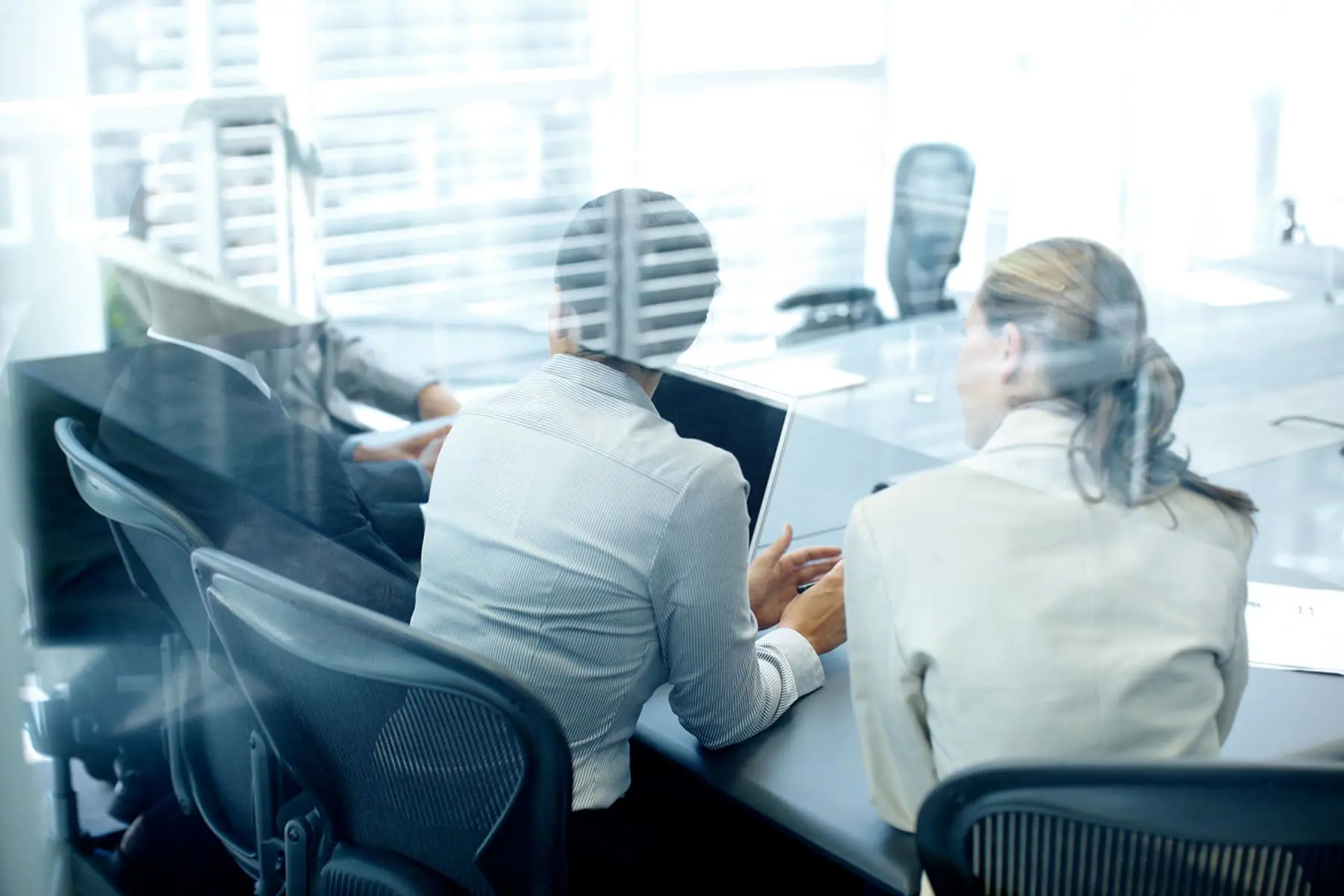 Two professionals are engaged in work at their computers in a modern office setting, with a view through a glass partition.
