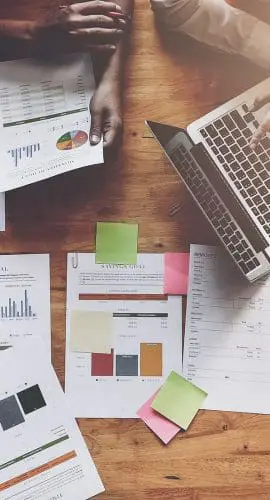A top view of a wooden table with a laptop, documents, sticky notes, and people's hands, suggesting a collaborative work environment.