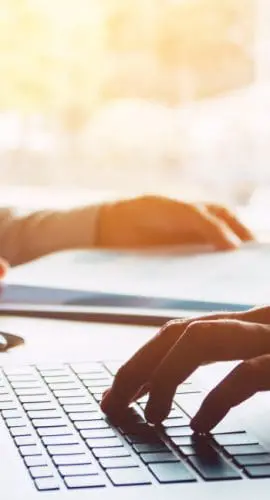 A person is typing on a laptop while another person points at the screen, suggesting a collaborative working environment.