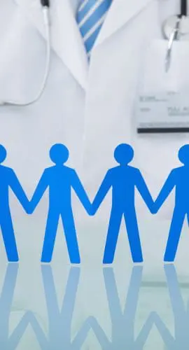 A person wearing a white lab coat and a stethoscope holds paper cutouts of blue human figures linking hands on a reflective surface.