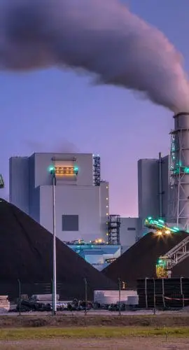 An industrial plant with a smoking chimney and conveyor belts is seen during twilight, with piles of black material in the foreground.