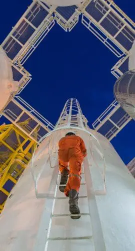 An individual in an orange suit ascends a ladder amidst towering white cylindrical structures against a twilight sky.