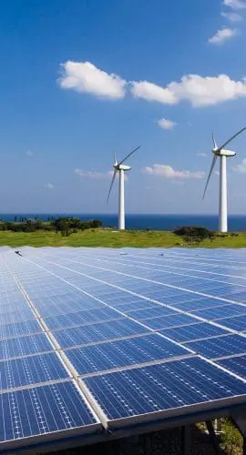 A solar panel array in the foreground with wind turbines in the background under a clear blue sky.