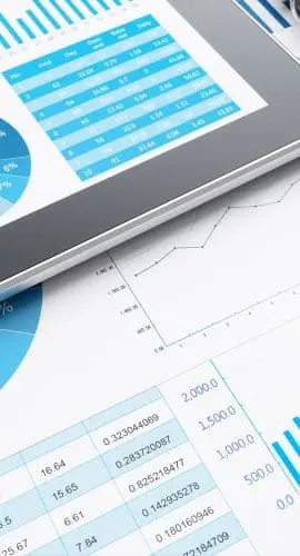 A tablet displaying colorful charts and graphs is lying next to printed data reports and a spiral-bound notebook on a white surface.