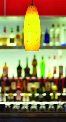 A warmly lit pendant lamp hangs above a bar counter lined with various bottles of alcohol against a red backdrop.
