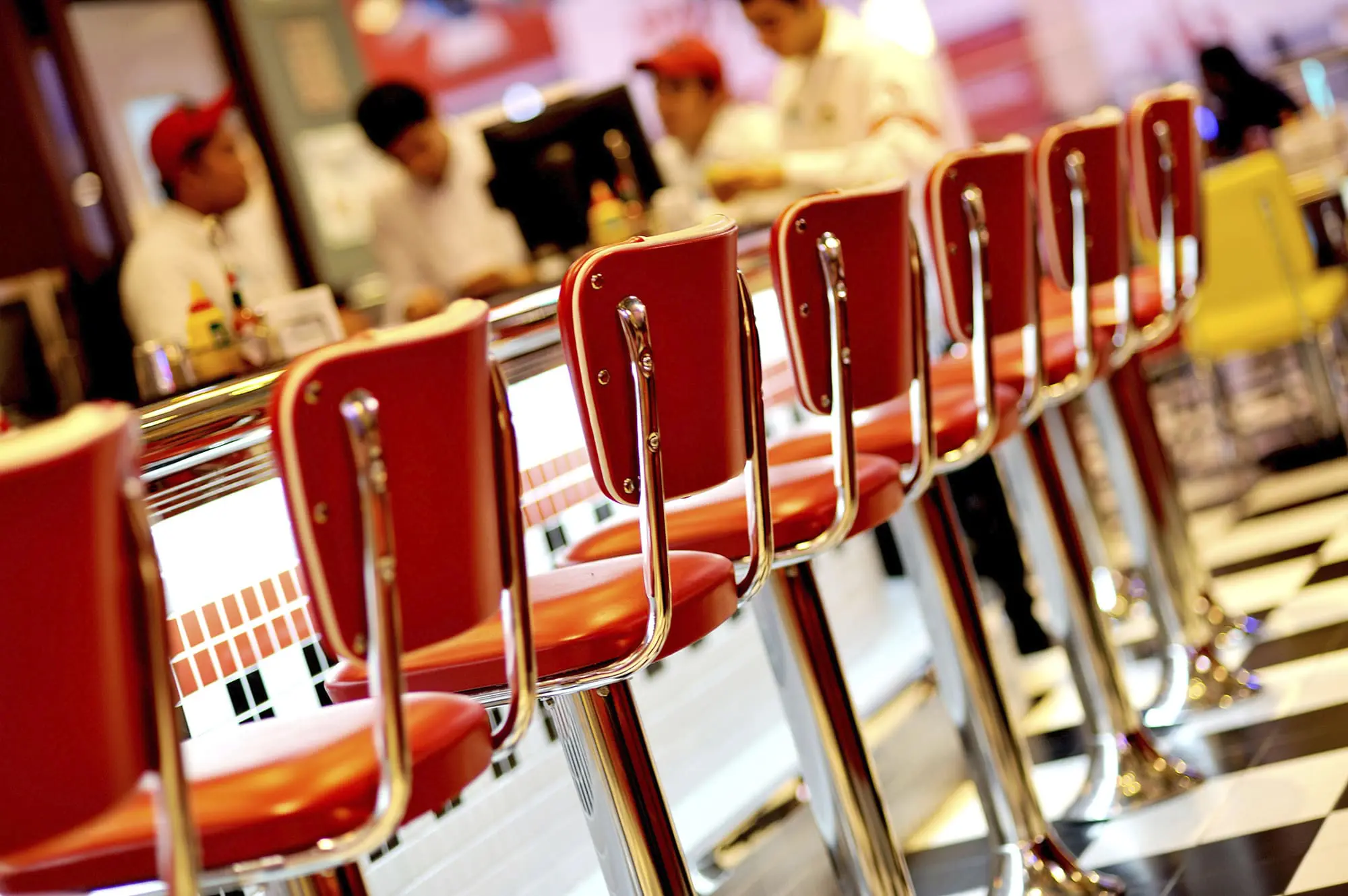 A row of red bar stools at a counter in a diner with staff in the background.