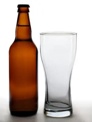 An empty brown beer bottle stands next to a clear pint glass against a neutral background.