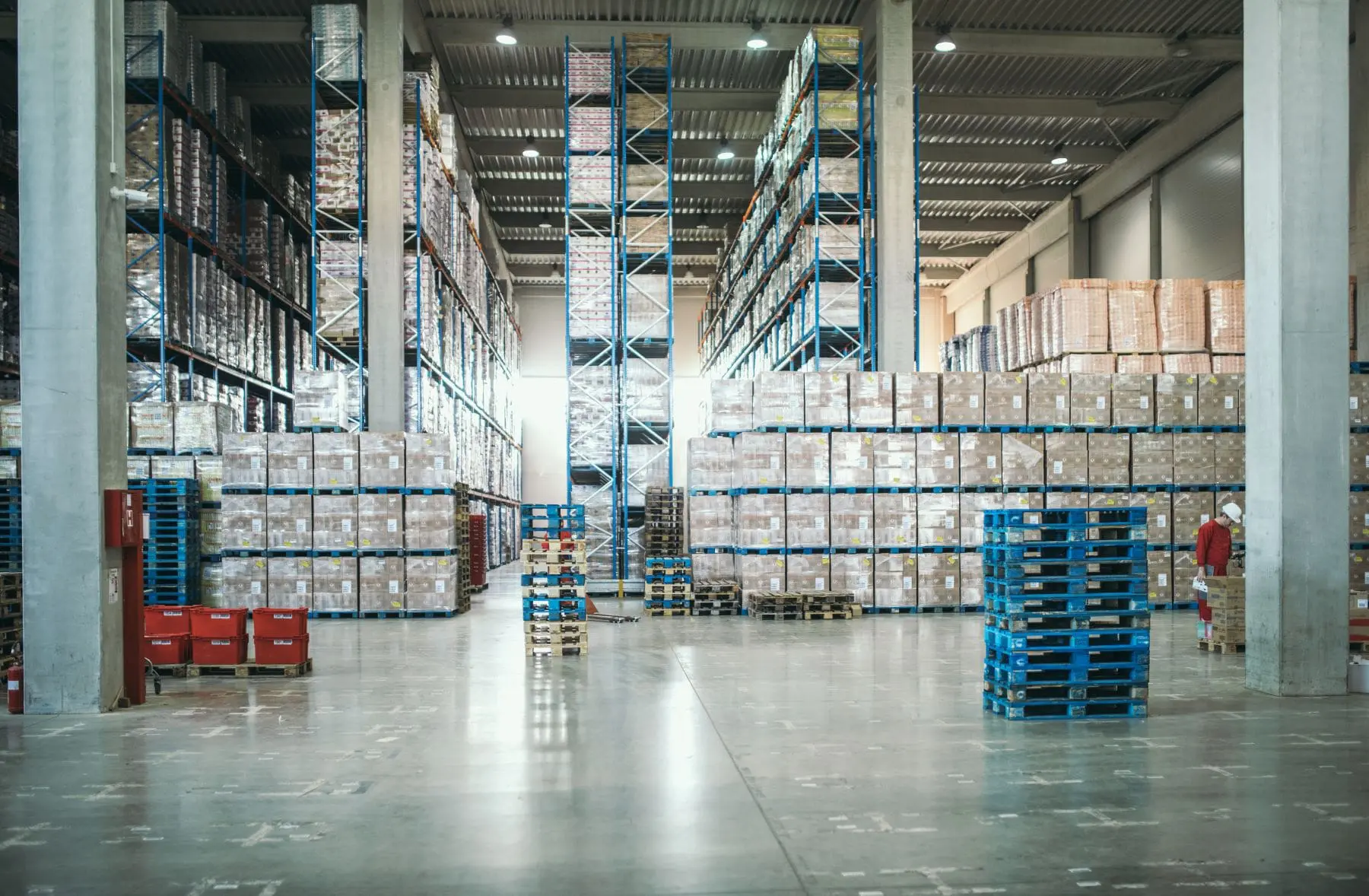 A spacious warehouse with high shelves stocked with numerous boxes and pallets arranged systematically on the floor.