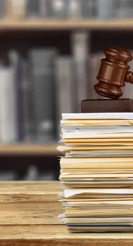 A wooden gavel rests on a stack of papers with a blurred backdrop of bookshelves.