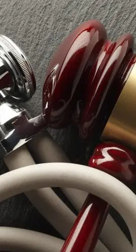 A close-up of three stethoscopes with silver chest pieces and tubes in shades of white and red, resting on a textured dark surface.
