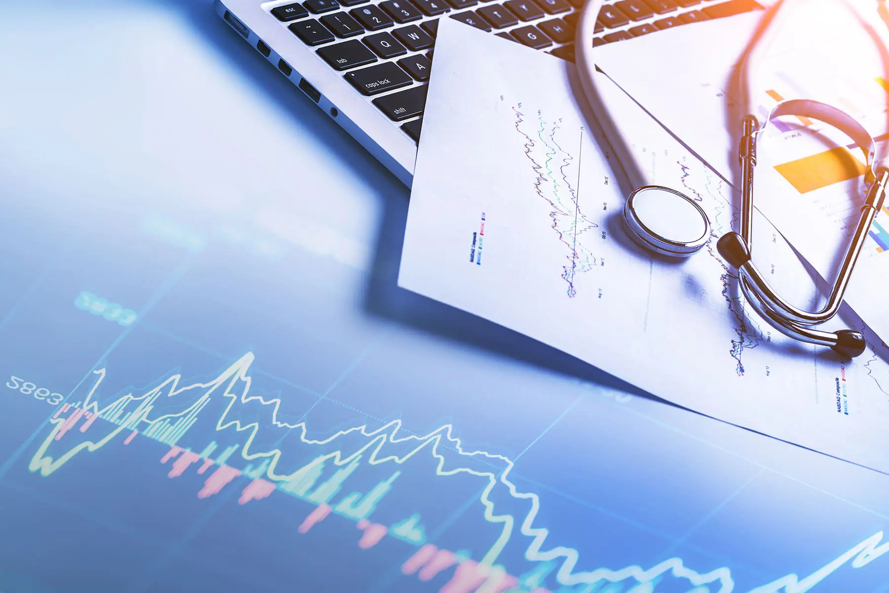 A stethoscope lies on top of documents beside an open laptop with medical and financial graphs visible in the background.