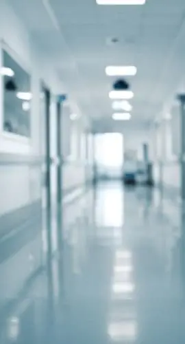 A blurry figure is visible at the end of a bright, clean hospital corridor with white walls and a reflective floor.