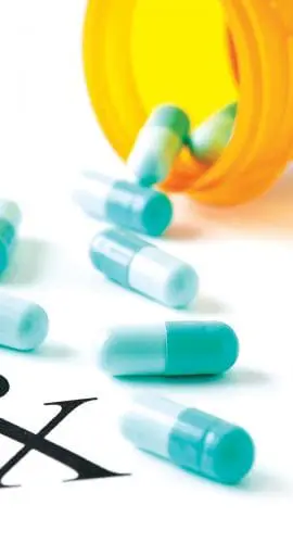 A prescription bottle lies on its side with blue and white capsules spilled out next to a bold "Rx" symbol on a white surface.