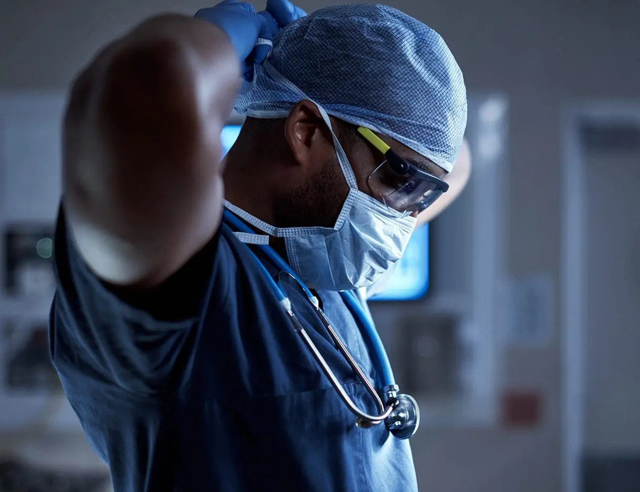 A healthcare professional is dressed in surgical scrubs, a mask, and protective eyewear while tying a cap behind their head.
