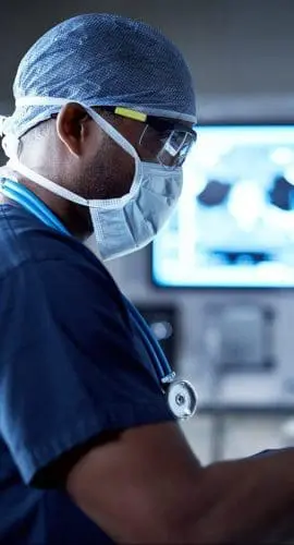 A healthcare professional in scrubs is focused on a task while monitoring information on a medical screen in a clinical setting.