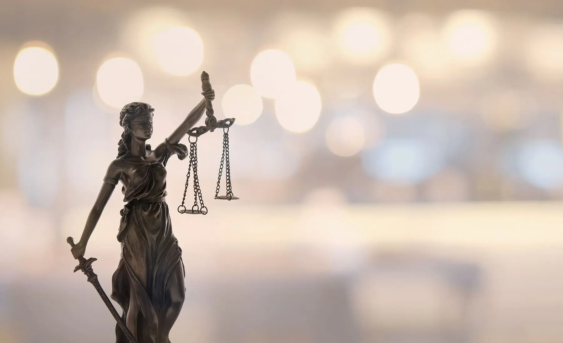 A statue of Lady Justice holding balanced scales symbolizes the fair administration of law, set against a bokeh light background.
