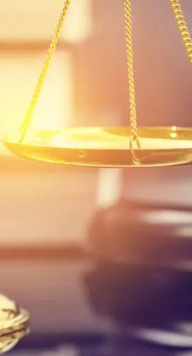 A set of golden scales is in focus in the foreground with a gavel and sound block blurred in the background, suggesting a legal or judicial setting.