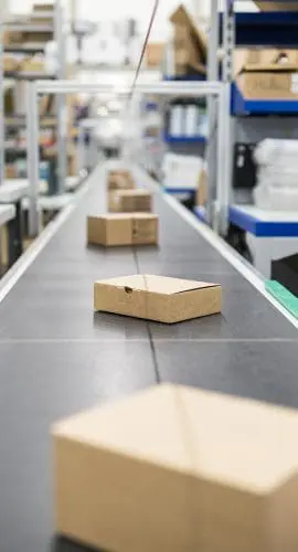 A conveyor belt in a warehouse carries cardboard boxes past shelves stocked with various items.