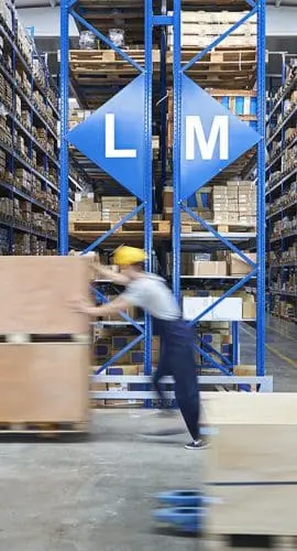 Workers in hard hats are actively moving goods around in a well-organized warehouse filled with tall shelving units.
